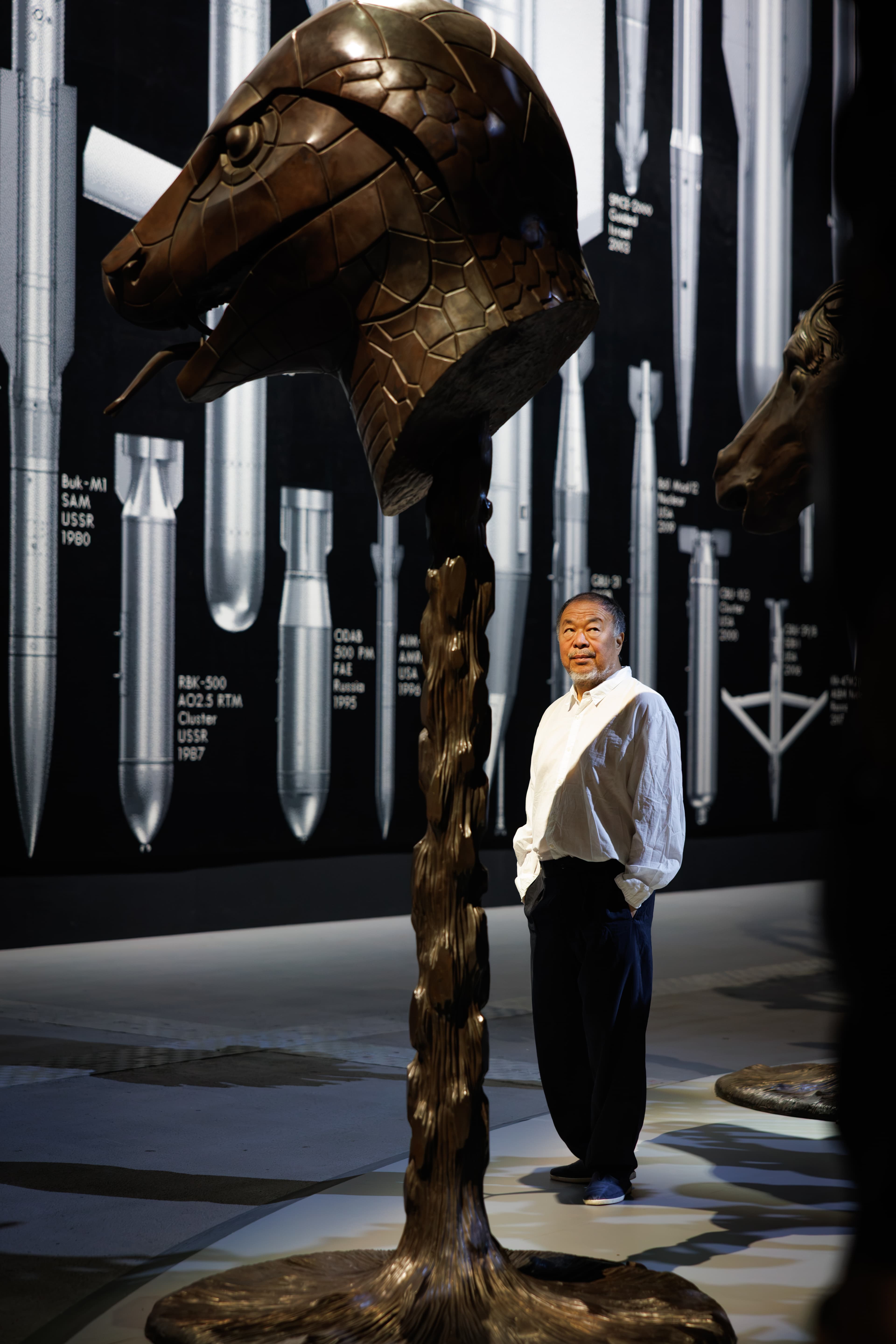 Ai Weiwei seated beneath a large sculpture inside a dark installation space.