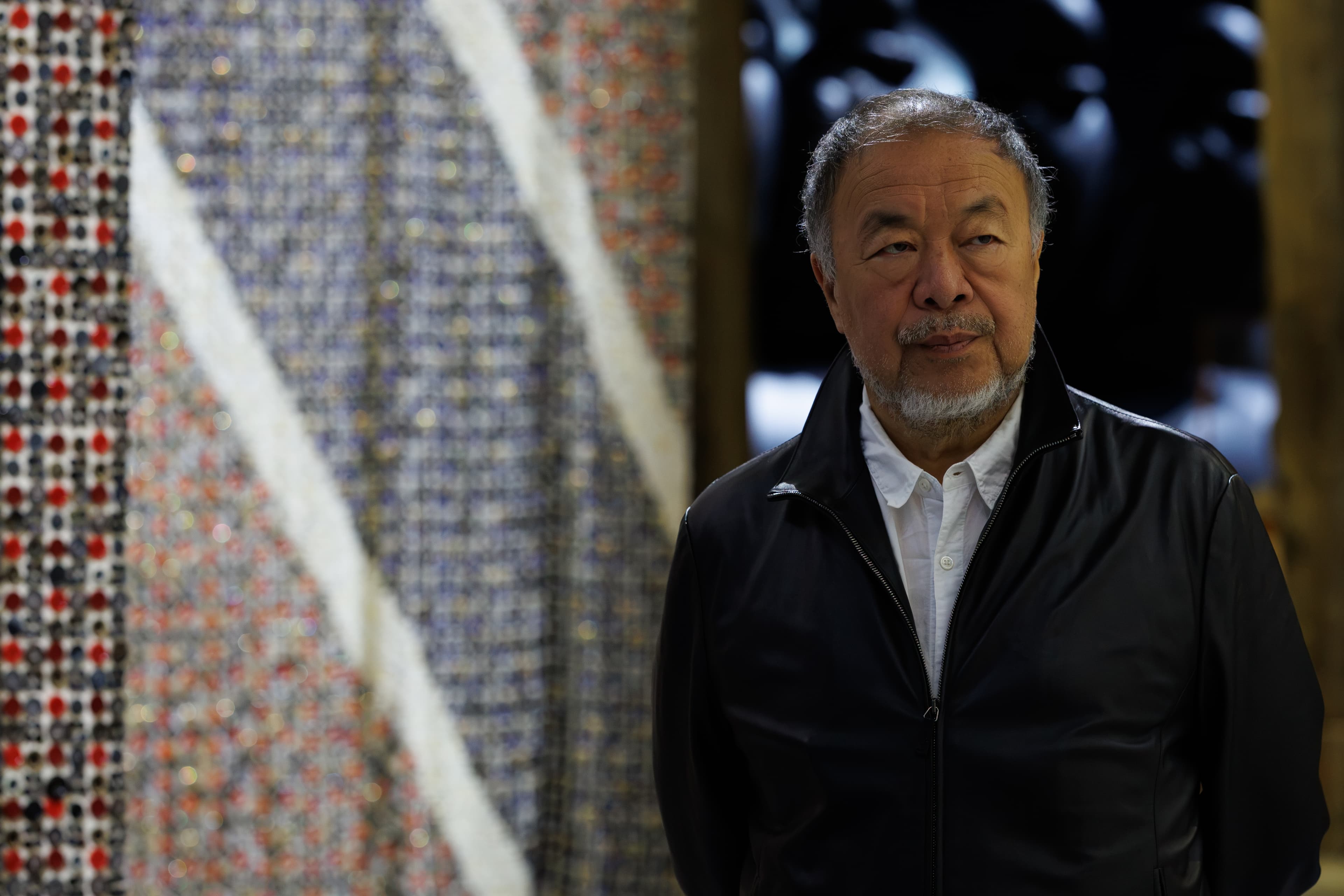 Ai Weiwei photographed in a dark coat in front of a textured installation at Factory International.