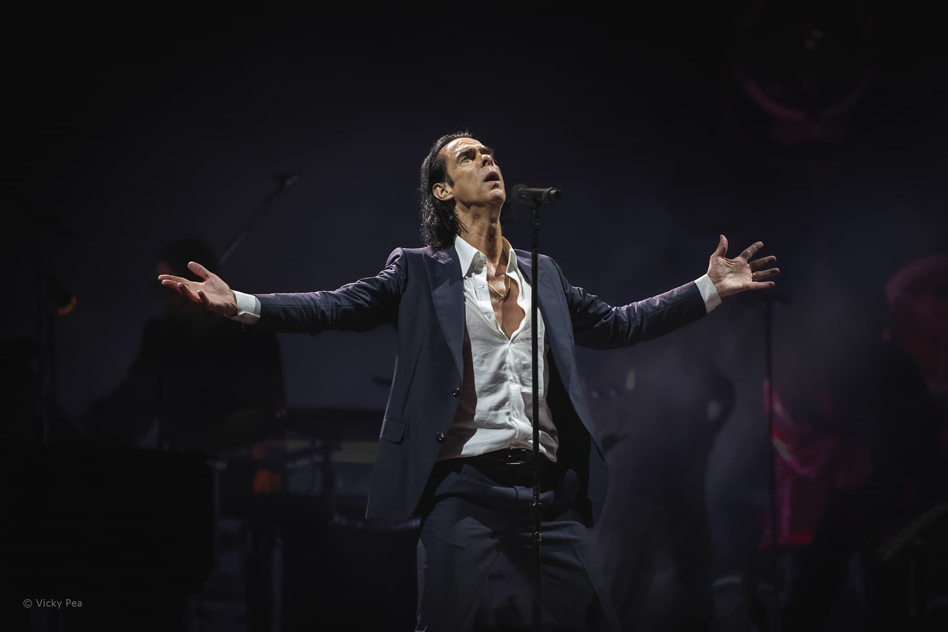Nick Cave performing at All Points East in London with arms open on a dark stage.